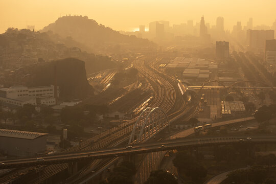 Aerial view of a complex railway junction and highway overpass with urban buildings and hills in the background under a warm golden sunset light in Rio de Janeiro, Rio de Janeiro, Brazil.