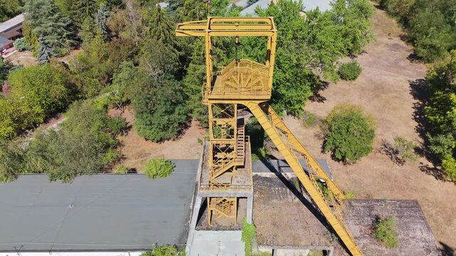 Aerial view of the historic Tiribesi-Akna coal mine headframe in B&aacute;tonyterenye, Hungary, standing above former industrial buildings amid summer trees and the M&aacute;tra foothills.
