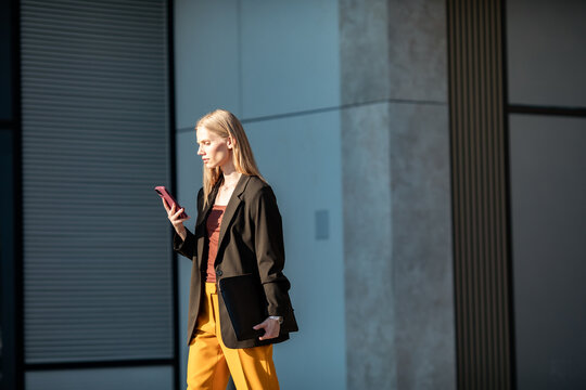 Businesswoman walking outside office, checking smartphone and carrying laptop