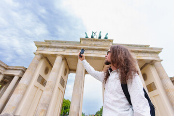 Long-haired traveler captures a digital memory using his smartphone while standing beneath the historic sandstone architecture of the famous Brandenburg Gate in Berlin. © Katie Chizhevskaya