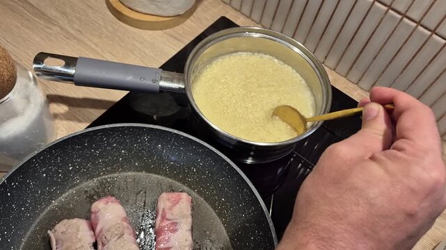 ​Close-up of a person stirring couscous with a spoon in a saucepan while frying bacon wrapped meat rolls in a pan on the stove, preparing dinner.