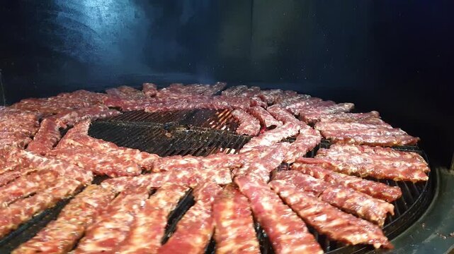 ​Raw juicy ribs or meat kebabs arranged in a circle on a large commercial BBQ rotisserie grill grate, preparing for roasting during a outdoor street food festival.