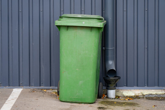Wheeled waste bin with a closed hinged lid stands beside a building downpipe, providing a movable refuse container for premises cleaning and routine waste collection