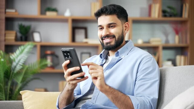 A happy smiling man uses a modern smartphone in a close up shot at home. He scrolls through a social media feed, chats cheerfully, and feels joyful while holding a wireless mobile device on a couch.