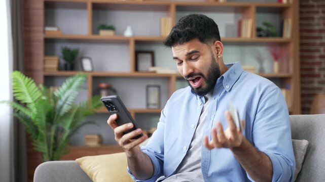 A shocked bearded man sitting on a sofa, looking at a smartphone screen and holding a credit card in frustration. A disappointed male experiences online banking fraud and money loss at home.