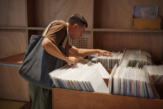 Vinyl record shopping in music store, man with canvas tote bag browsing album bins, searching for LP music and enjoying retro analog hobby during leisurely shopping trip.