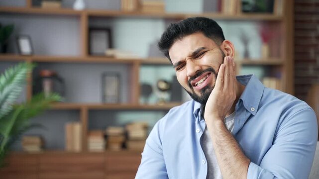 Close up of man suffering from severe toothache, holding hand near cheek. Unhappy person touching sore area on swollen face, experiencing intense dental pain, stress, and feeling unwell in room.