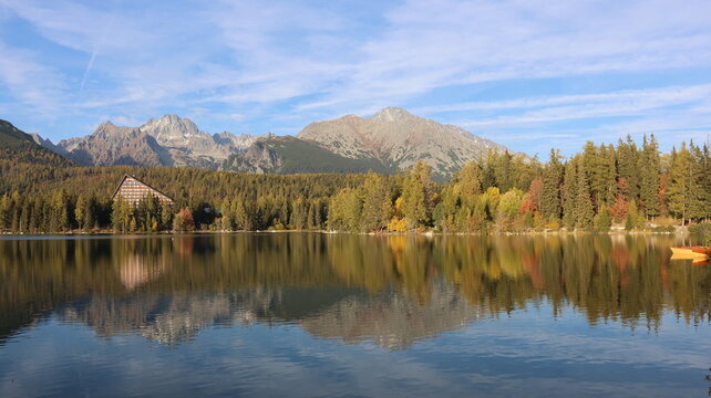 Scenic Mountain Lake with Autumn Forest Reflections in the High Tatras, Slovakia. The calm water reflects golden trees, rugged mountains, and a blue sky, creating a peaceful natural landscape 
