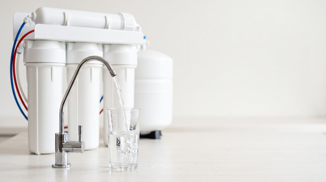 Modern reverse osmosis water filtration system purifying liquid into transparent glass on clean kitchen counter against bright wall background closeup with copy space representing clean drinking