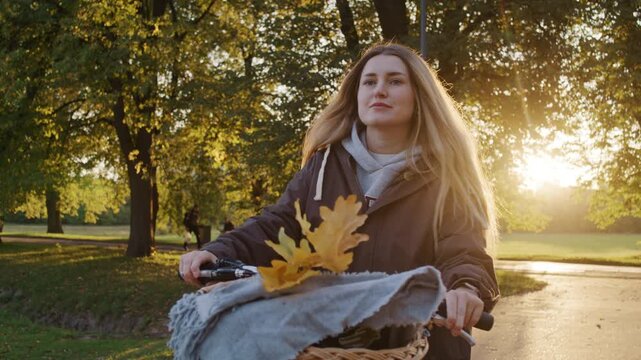 Cheerful woman cycles past tall trees in sunset autumn park