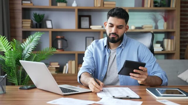 A focused man sits at his home office desk using a calculator while checking financial data on his laptop. He carefully fills out a paper form and writes detailed notes to manage his family budget.
