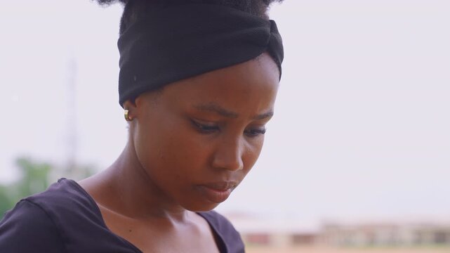Close up portrait captures young woman with dark skin wearing black headwrap. Woman maintains steady eye contact downwards revealing minimal facial expression variation