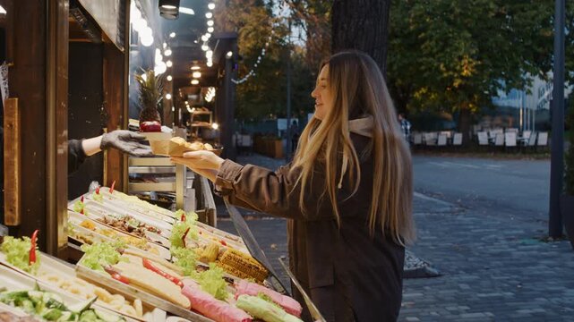 Pretty woman buys fast food in street kiosk in evening city