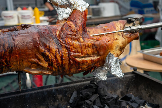 Whole Roasted Pig on Metal Rotisserie Spit with Crispy Golden Brown Skin Over Charcoal Grill at Outdoor Food Market