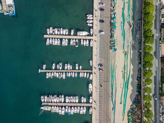 Aerial view of a busy mediterranean marina and concrete forecourt with bright green fishing nets drying by piers, coastal road and seaside houses on a sunny day © Osvaldo Mussi