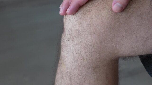 A man grips and massages his painful knee with both hands, the bent hairy leg seen from the side against a plain grey background. A close up for knee pain, joint ache, arthritis, sports injury and ph