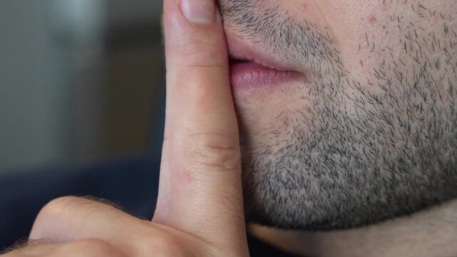 A man with a stubble beard raises his index finger to his lips in a shush gesture, his face cropped from the nose down against a blurred background. A close up for silence please, keep a secret, quie