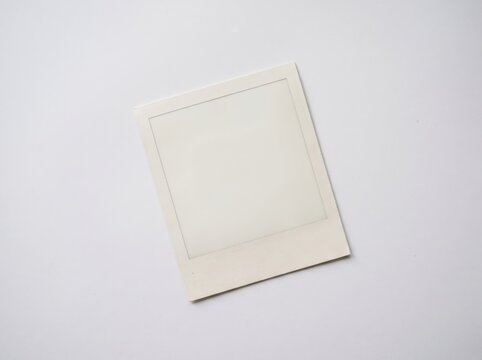 Blank instant photo frame tilted on white background, empty photo template. A slightly rotated square print with a wide white bottom border and an empty white center rests on a plain white surface