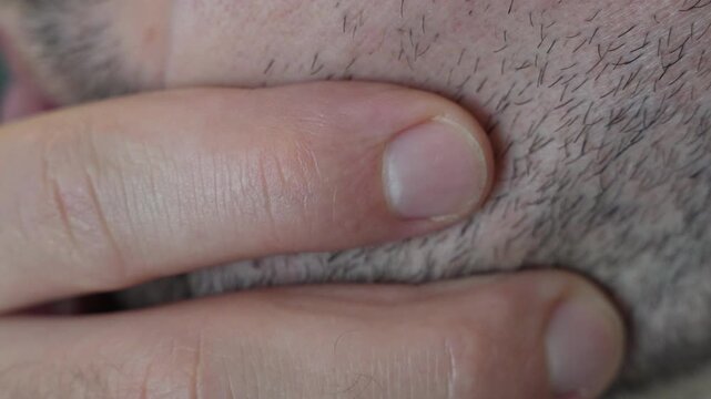 A man&rsquo;s thumb strokes along his stubbled chin below his lips, filmed in tight macro with every coarse hair and pore visible. A detail shot for beard growth, rough stubble, a decision to shave, a thin