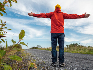 Person stands with arms wide open on gravel road. Red jacket and yellow hat contrast with blue sky. Feels freedom, peace, or adventure. Great for travel, wellness, or outdoor lifestyle themes. © mark_gusev