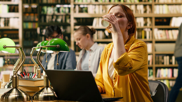 Coffee, study break and laptop as woman sips a takeaway drink beside her computer among other readers. Campus life, refreshment and independent learning accompany coursework in a library.