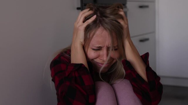 Young blonde woman sits rocking and holding her head, conveying anxiety, emotional instability, severe distress and a nervous breakdown