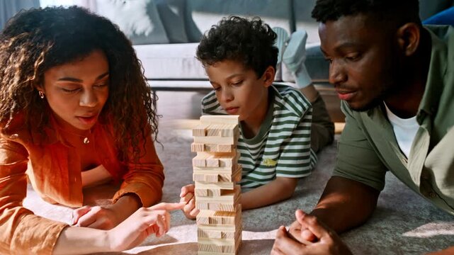 Multiethnic family playing wooden block tower game. Parents son lying carpet.