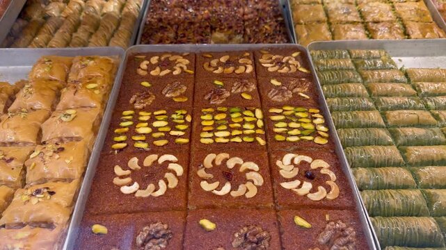 Traditional Turkish baklava desserts with pistachios, walnuts, and cashews in metal trays inside a bakery shop window. Oriental sweet pastry, middle eastern confectionery, gourmet dessert variety.
