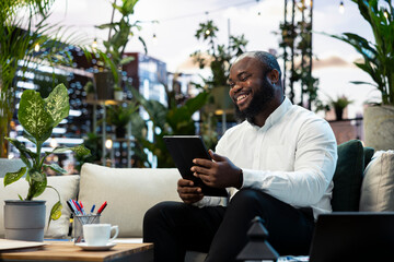 Businessman, tablet and technology as smiling professional reads handheld screen while relaxing on sofa. Productivity, research and digital workflow support independent work in urban rooftop lounge. © DC Studio