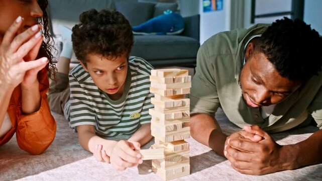 Boy pulling wooden block tower parents watching. Family playing floor game home.