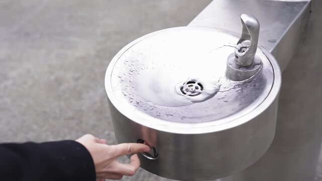 Hand pressing button on public drinking water fountain outdoors, fresh clear water stream pouring from metal bubbler faucet concept.