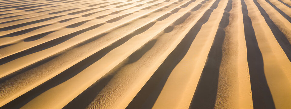 Rippled sand dune surface with parallel diagonal ridges and shadows, golden sand seen from above.