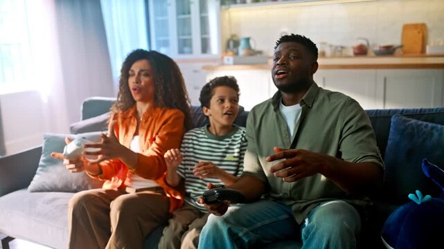 Son comforts disappointed father after video game loss. Mom laughing on sofa.