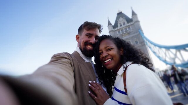 A Latina African American woman and a Caucasian man smile in a handheld selfie on Tower Bridge in London, England. They lean together, turn towards one another and share a kiss.