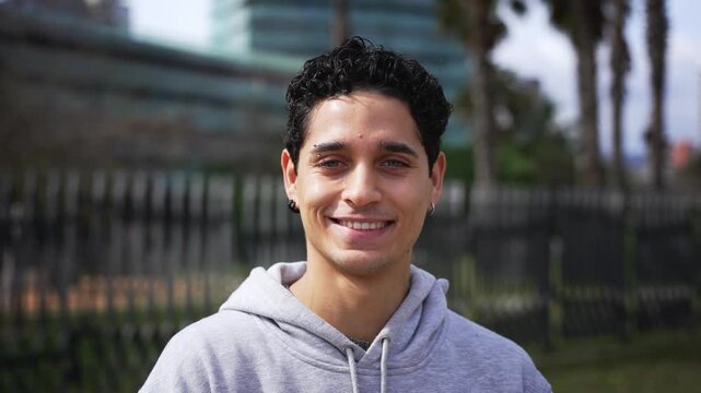 A young Latino Venezuelan man with short curly hair and a gray hoodie smiles towards the camera in a park. A fence, palm trees and buildings form the background as he gently tilts his head.