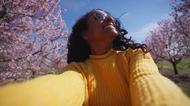 A Latina African American woman in a yellow sweater and sunglasses films a playful selfie among rows of pink flowering almond trees. She smiles, raises arm and look the camera while walking through