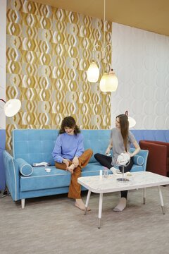 Two young women are engaged in skin care procedures at home sitting on a cozy sofa. One woman rubs the cream in the feet