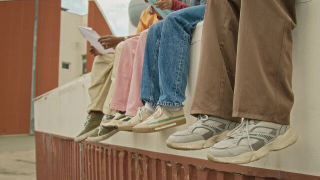 Multiethnic teenagers dangling legs while sitting on concrete wall outdoors. Footage suitable for youth lifestyle campaigns, footwear advertising and student social life promotions