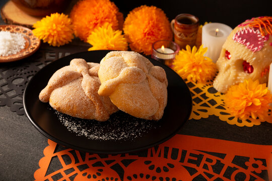 Traditional Mexican ofrenda, Pan de Muerto sweet bread on a black plate, placed on a Day of the Dead altar setup with cempasuchil marigold flowers and sea salt background.