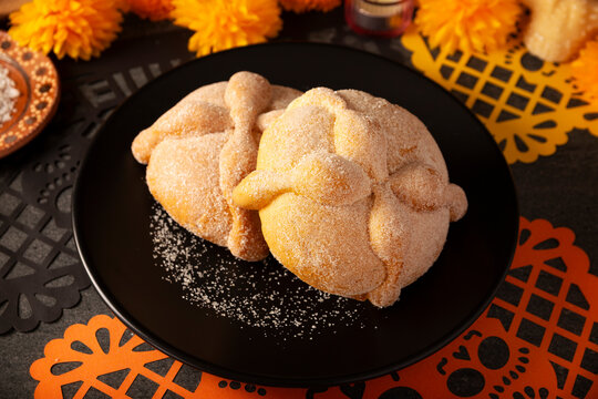 Mexican Pan de Muerto sweet breads on a matte black plate next to a festive orange papel picado paper cutout for Day of the Dead Ofrenda.