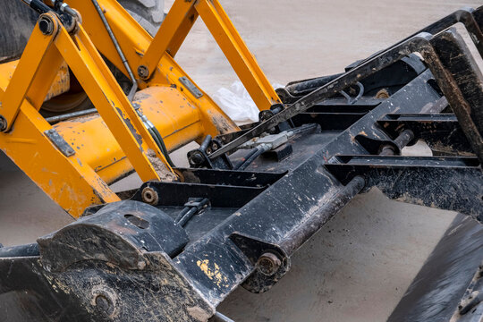 Backhoe loader bucket close-up. Worn yellow frame and black steel bucket with hydraulic hoses and pivot bolts fill the frame. Suits construction, engineering, and infrastructure content.