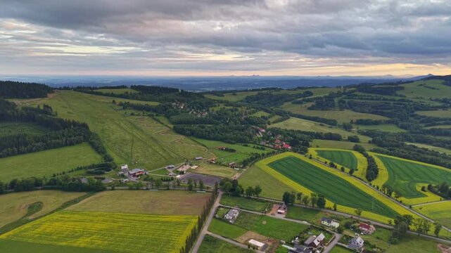 From high above, patchwork fields and rolling green hills stretch toward small village of houses and winding roads, with warm sunset light, wide horizons.