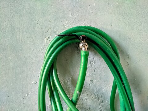 Green water garden hose coiled around metal tap faucet on textured wall