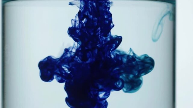 Deep cobalt blue and teal ink billow and diffuse through clear water, forming intricate organic clouds against a pale cyan background. The fluid motion creates dramatic contrast, delicate tendrils, an