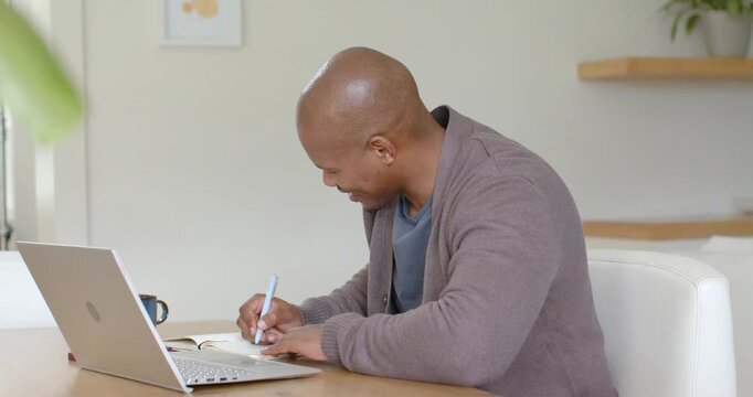 African American man leaning over laptop, reading screen, writing notebook, smiling