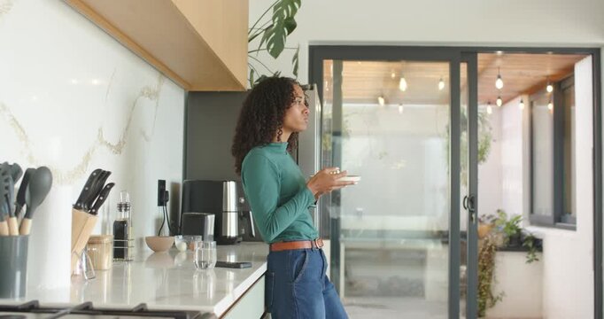 African American woman in teal top leaning in kitchen sipping cup holding saucer glancing at patio