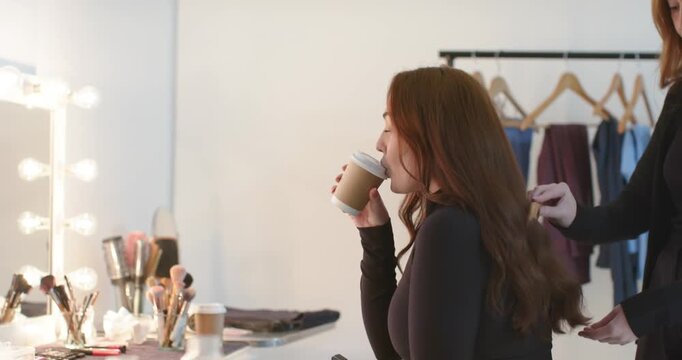 Stylist combing hair with wide-tooth comb as client lifting paper cup wearing black top at vanity