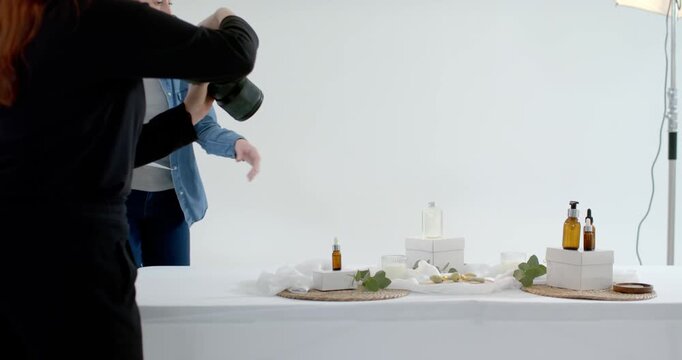 Female colleagues stylist entering from right adjusting clear bottle on box risers, aligning frame