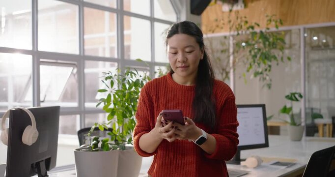 Adult Asian woman sitting on desk, glancing at window then checking messages on smartphone, smiling