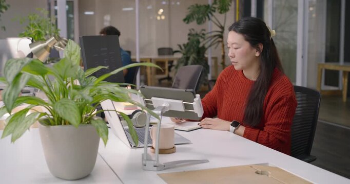 Asian woman wearing rust sweater at office shifting between tablet on stand and laptop